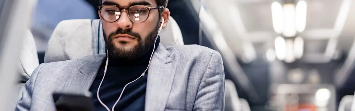 A man sitting, focused on his phone, wearing glasses and earphones, dressed in a blazer.