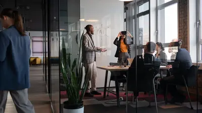 Ein modernes Büro mit großen Fenstern und Glaswänden. Im Raum stehen mehrere Personen, die an einem Tisch sitzen und an Laptops arbeiten. Ein Mann steht vor einer weißen Tafel und erklärt etwas, während eine Frau daneben eine VR Brille testet. Im Vordergrund ist eine große Pflanze zu sehen.