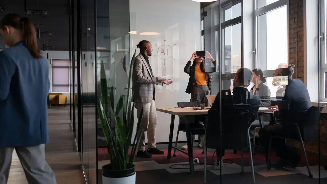 Ein modernes Büro mit großen Fenstern und Glaswänden. Im Raum stehen mehrere Personen, die an einem Tisch sitzen und an Laptops arbeiten. Ein Mann steht vor einer weißen Tafel und erklärt etwas, während eine Frau daneben eine VR Brille testet. Im Vordergrund ist eine große Pflanze zu sehen.
