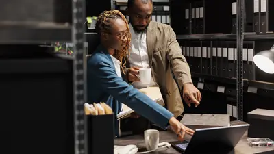 Two people in an archive room, filled will folders, pointing to the laptop screen