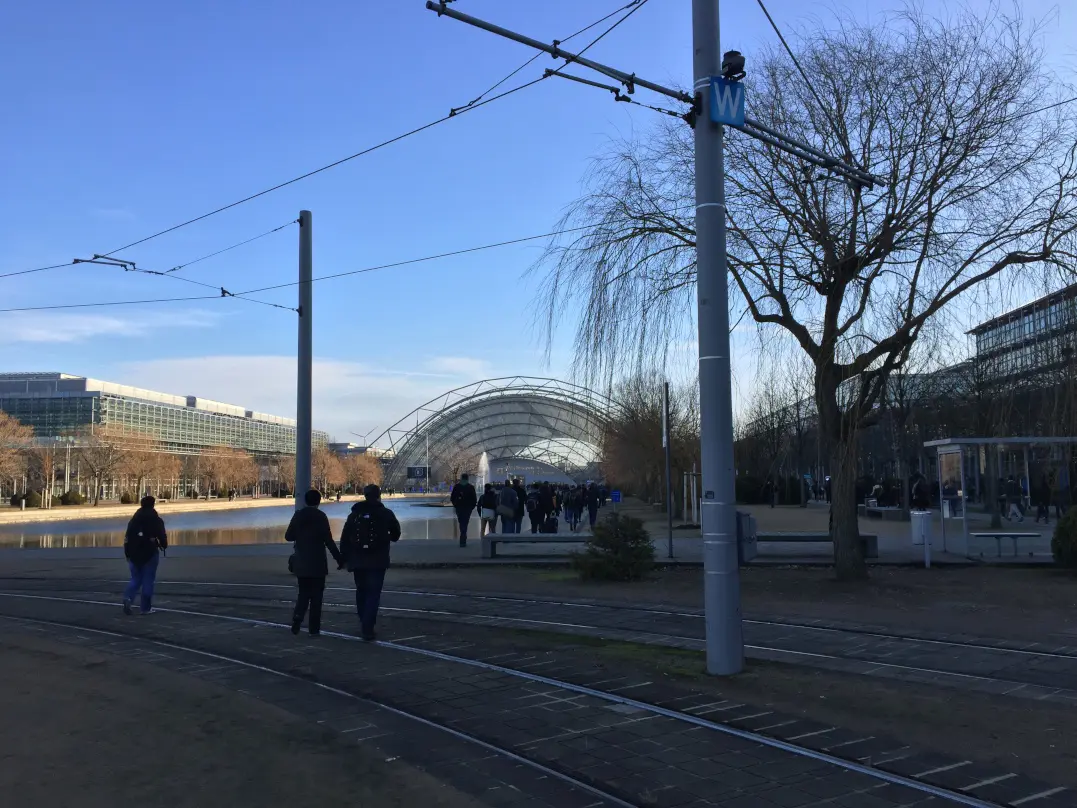 Blick auf eine Straßenbahn, die auf einer Schiene fährt, mit einem blauen Himmel im Hintergrund. Es sind einige Passanten zu sehen, die den Bahnsteig entlanggehen.
