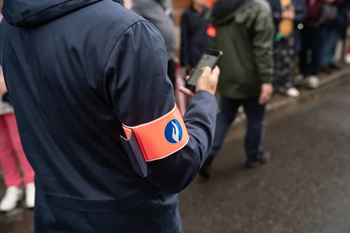 Person wearing a dark jacket with a red armband, standing among a crowd, possibly at an event or protest.