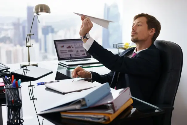 A  man sitting at a cluttered office desk, holding a paper airplane and looking thoughtfully into the distance.
