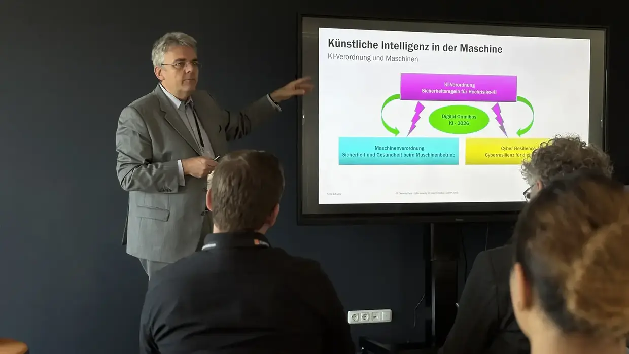Ein Mann hält einen Vortrag vor einer Gruppe. Er zeigt auf einen großen Bildschirm, auf dem eine Präsentation mit Diagrammen und Text zu sehen ist.