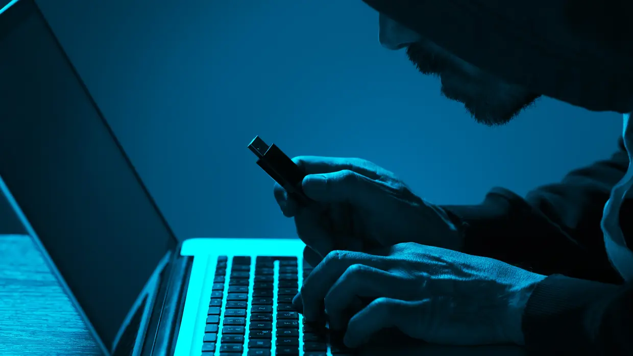 Een man steelt gegevens van een laptop in een donkere kamer en houdt een usb-stick in zijn hand.