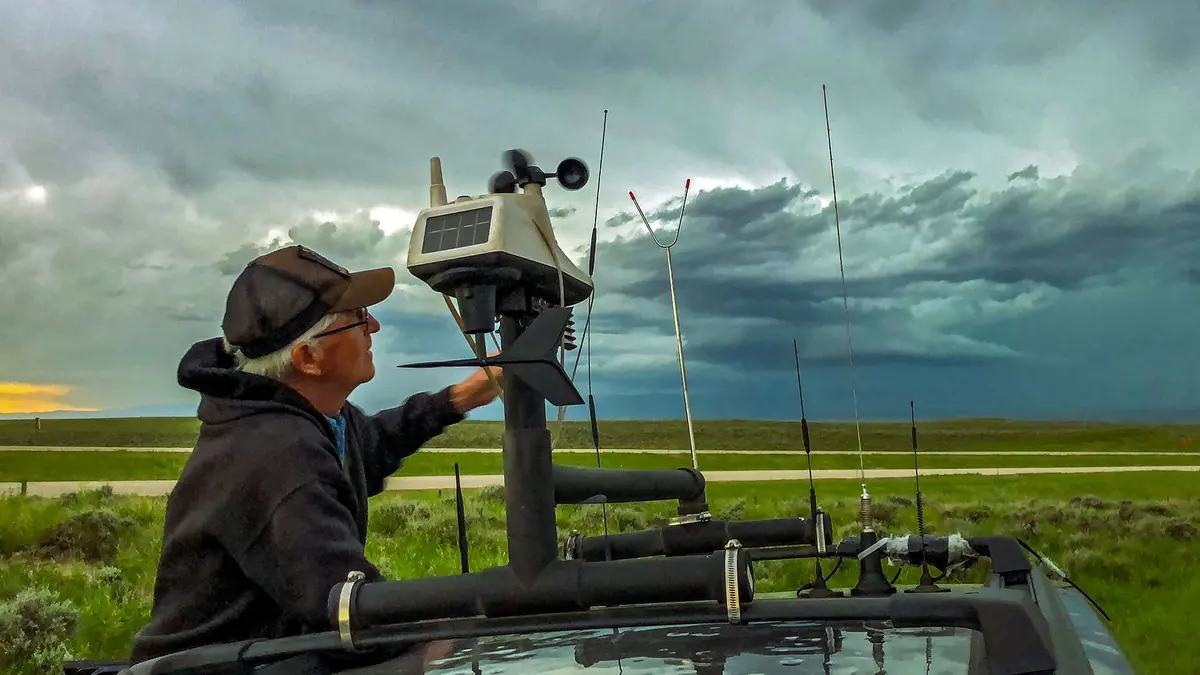 A person in outdoor gear adjusting a weather monitoring device on a vehicle, with stormy clouds and open fields in the background.
