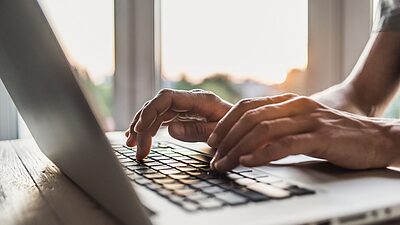 A close-up of hands typing on a laptop keyboard, with a bright window in the background.