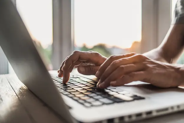 A close-up of hands typing on a laptop keyboard, with a bright window in the background.