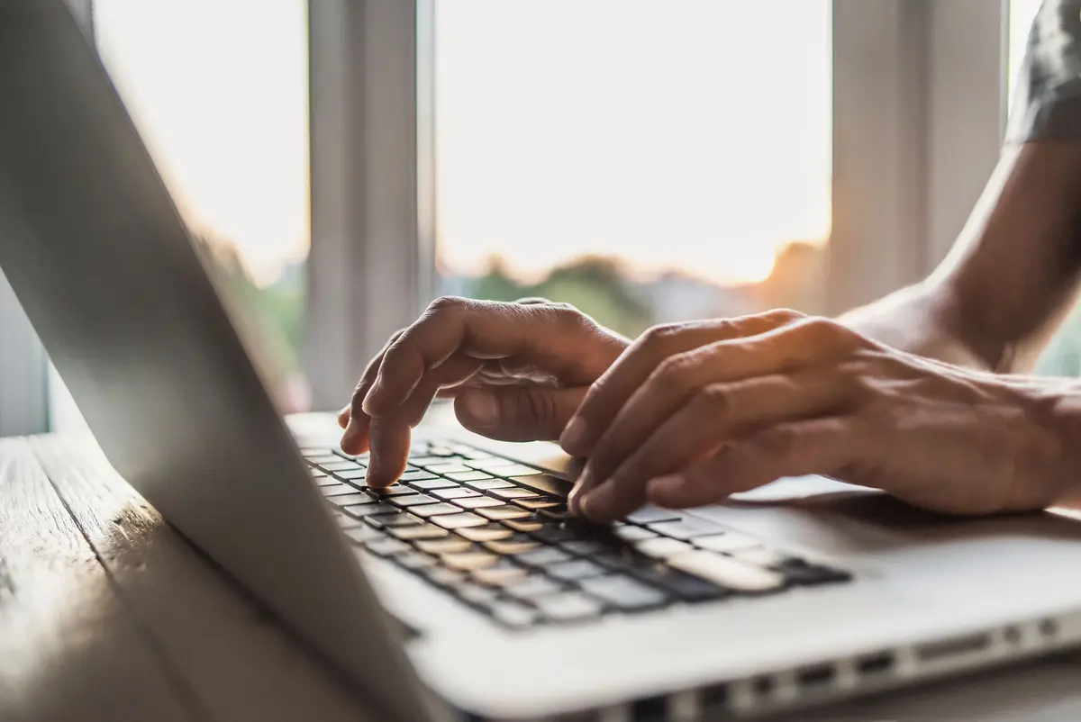 A close-up of hands typing on a laptop keyboard, with a bright window in the background.