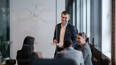 Ein Mann in einem dunklen Anzug steht in einem modernen Konferenzraum vor einer weißen Tafel mit einer Skizze zum Thema „AI“ (Künstliche Intelligenz). Er hält Papiere in der Hand und lächelt, während vier Frauen im Sitzkreis ihm zuhören und lachen. Der Raum ist gut beleuchtet mit großen Fenstern im Hintergrund.