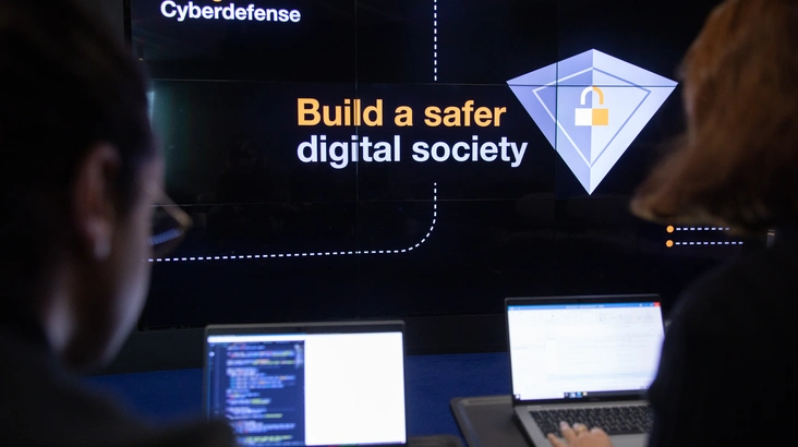 Zwei Personen arbeiten an Code, im Hintergrund steht "Build a saferdigital society"