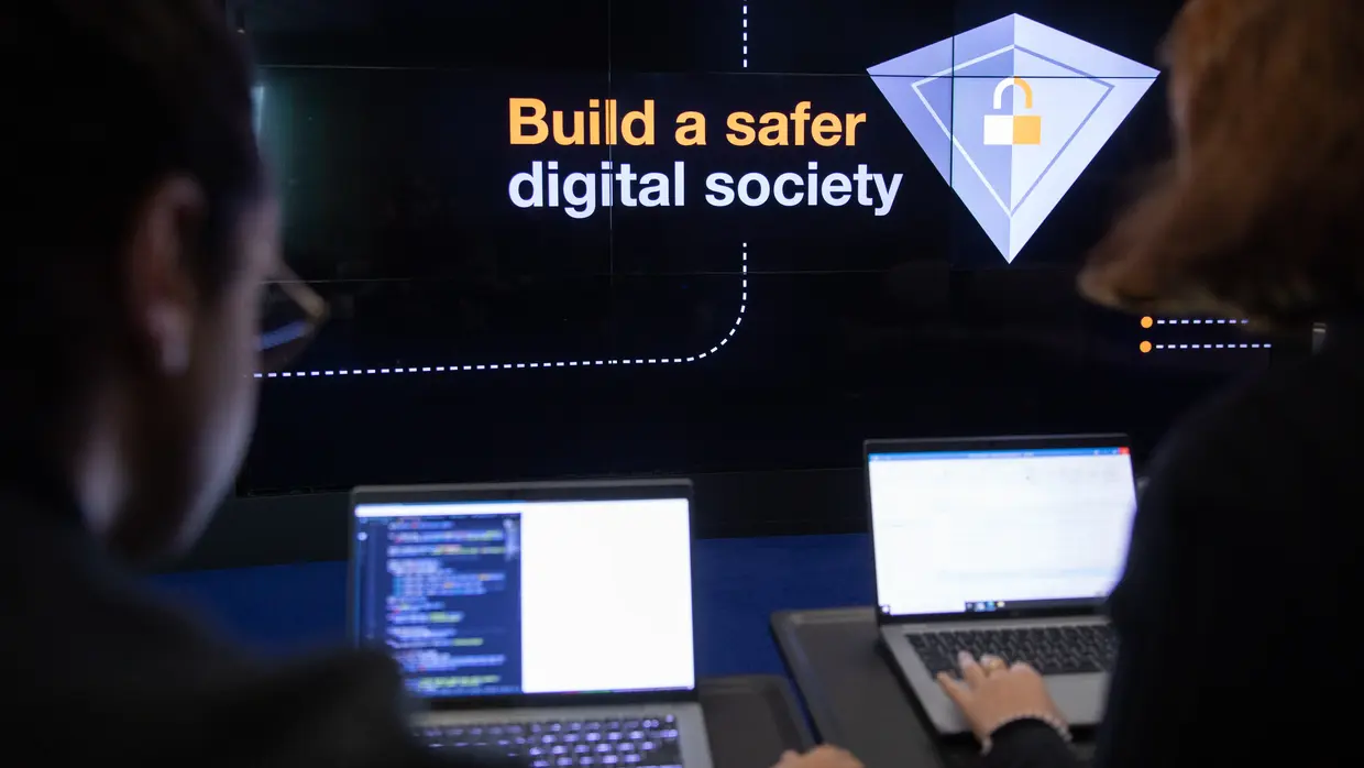 Zwei Personen arbeiten an Code, im Hintergrund steht "Build a saferdigital society"