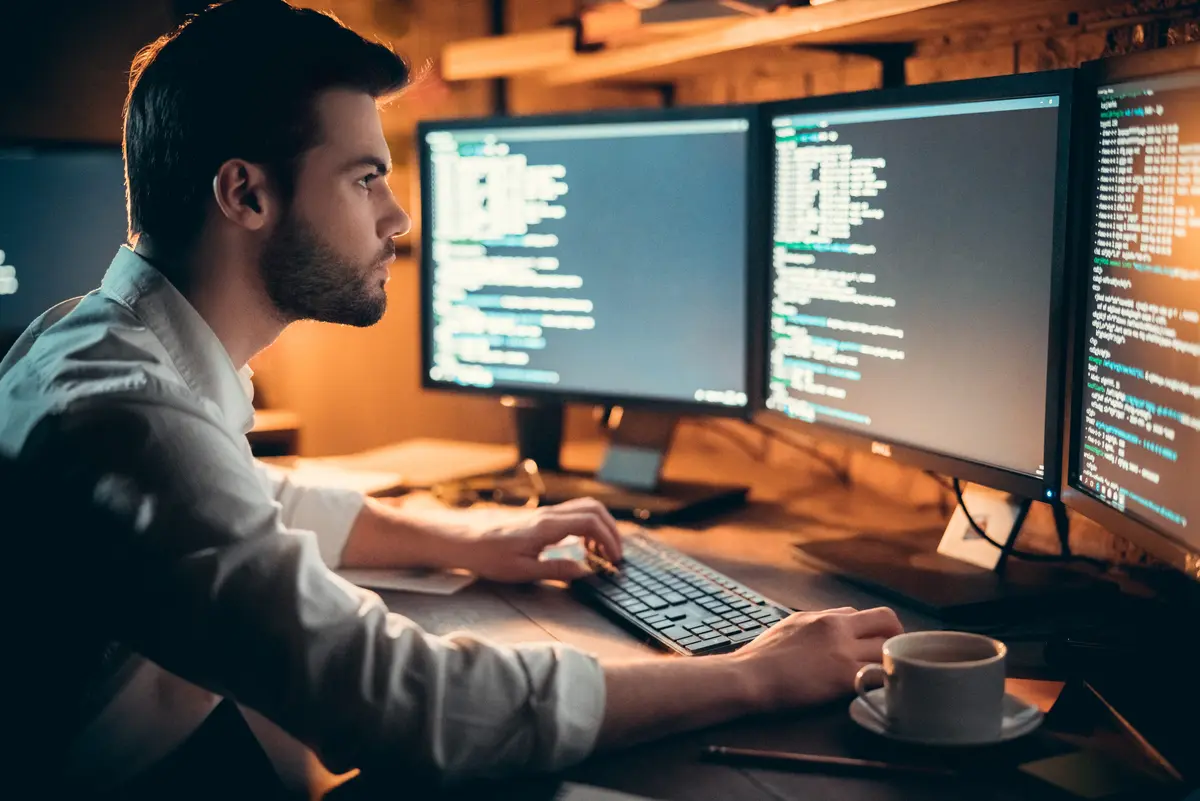 A man working intensely at a desk with multiple monitors displaying lines of code, highlighting a programming or cybersecurity environment.