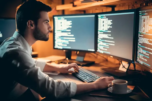 A man working intensely at a desk with multiple monitors displaying lines of code, highlighting a programming or cybersecurity environment.