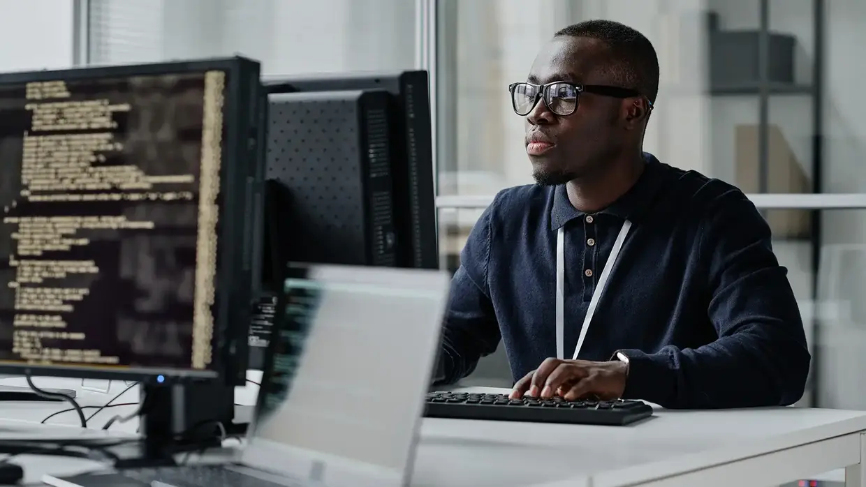 Ein Mann mit Brille sitzt an einem Schreibtisch vor mehreren Computermonitoren, arbeitet an einem Programm.