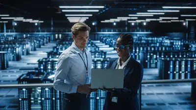 Two people holding a laptop with a server farm behind them