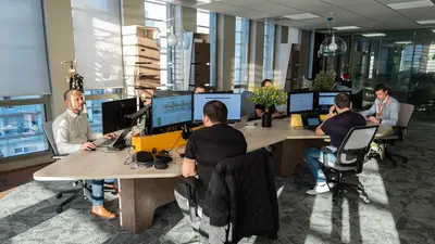Six hommes travaillent dans un open space lumineux, chacun devant un ordinateur portable relié à deux écrans.