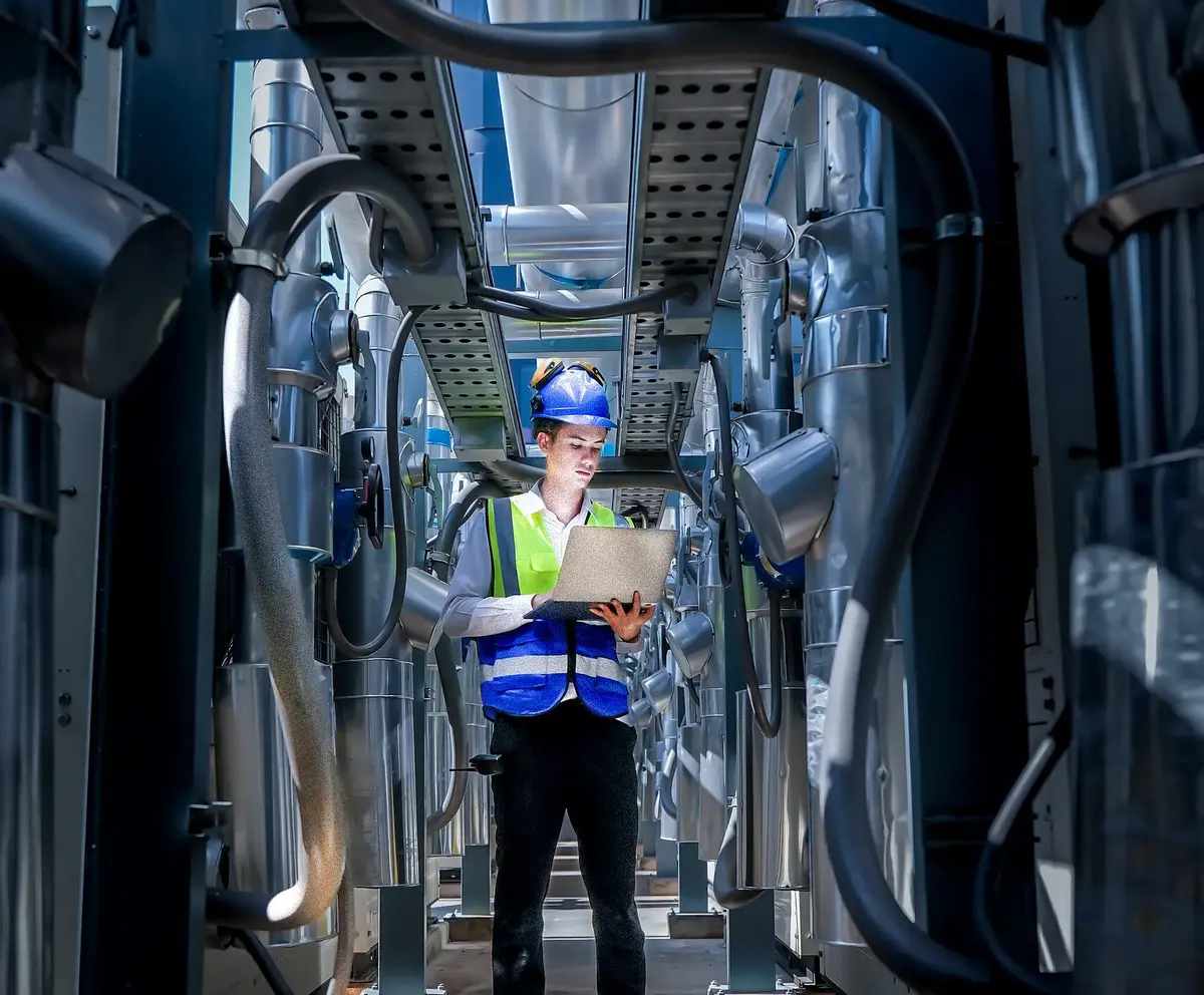 An industrial worker in high-visibility gear and a blue helmet meticulously examines the intricate network of machinery. The engineer is dedicated to maintaining operational efficiency and safety.