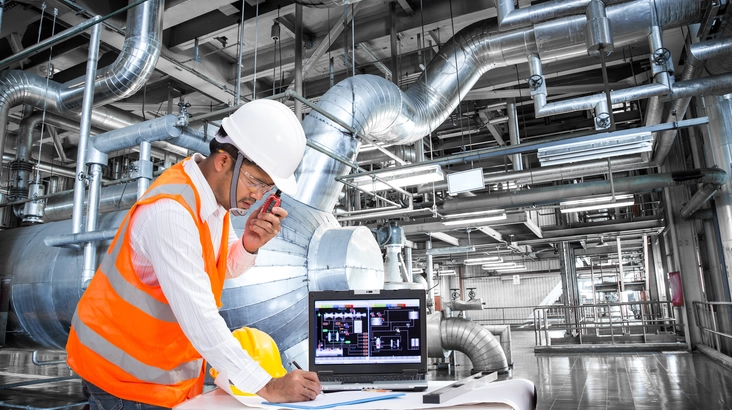Elektroingenieur mit Brille und Schutzhelm, Warnweste und Walkie-Talkie bei der Arbeit im Kontrollraum eines modernen Wärmekraftwerks.