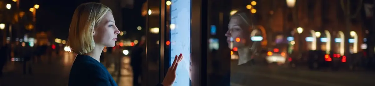 Une femme utilise un écran tactile en plein air la nuit, avec des lumières de ville en arrière-plan.