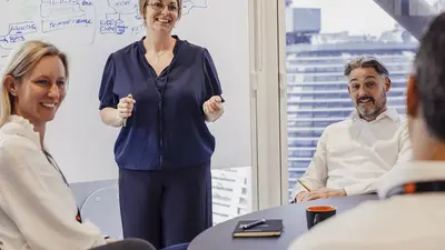 Eine Frau steht vor einer Gruppe in einem Meetingraum, lächelt und gestikuliert, während die anderen drei Personen am Tisch zuhören.