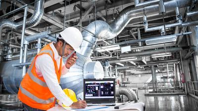 Ingenieur in einem weißen Schutzhelm und orangefarbener Sicherheitsweste arbeitet in einer industriellen Anlage mit zahlreichen Rohren und Ventilatoren. Er benutzt ein Tablet, um Daten zu überwachen oder zu steuern.