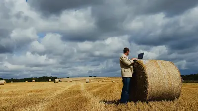 Ein Mann steht in einem Getreidefeld neben einem großen, runden Heuballen und arbeitet an einem Laptop. Der Himmel ist bedeckt mit dunklen, wolkigen Wolken.