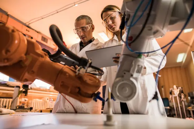 Two scientists or engineers working together in a laboratory or industrial setting, examining a robotic arm or automated machinery.