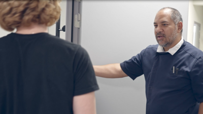 A photo of two people in a professional setting, with one person (facing away) wearing a dark shirt and the other (facing forward) in a lighter shirt, engaged in a handshake or conversation.
