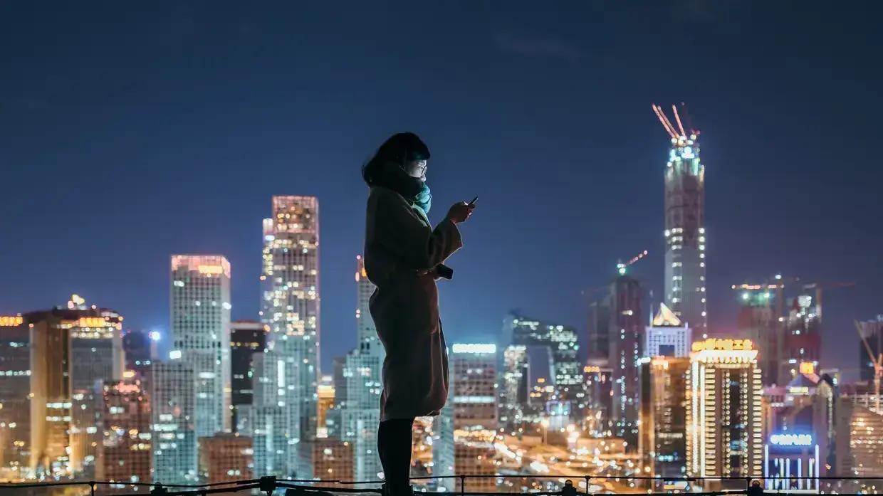 Eine Frau steht auf einem Dach vor einer nächtlichen Skyline mit Wolkenkratzern.