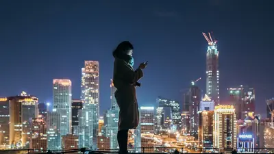 Eine Frau steht auf einem Dach vor einer nächtlichen Skyline mit Wolkenkratzern.