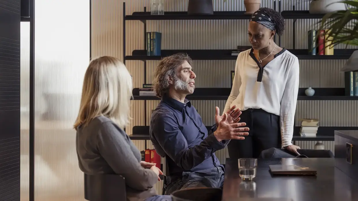 Drei Personen in einem Büro, im Gespräch. Ein Mann spricht, zwei Frauen hören zu.