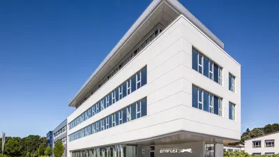 Ein modernes, weißes Bürogebäude mit mehreren Etagen und großen Fenstern, das unter einem klaren blauen Himmel steht.