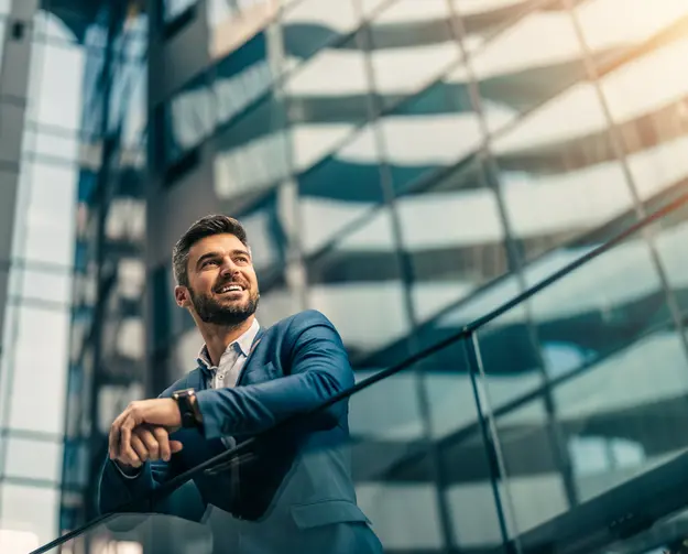 Smiling businessman leaning on a railing in a modern glass building, looking ahead with confidence