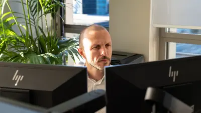 Un homme est assis dans un bureau, face à deux ordinateurs posés devant lui. Sa tête est tournée vers l’écran de droite.