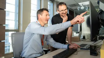 Deux hommes sont dans un bureau. L’un est assis devant un ordinateur et en désigne certains éléments du doigt, tandis que l’autre, debout à ses côtés, observe attentivement ce qu’il lui montre.
