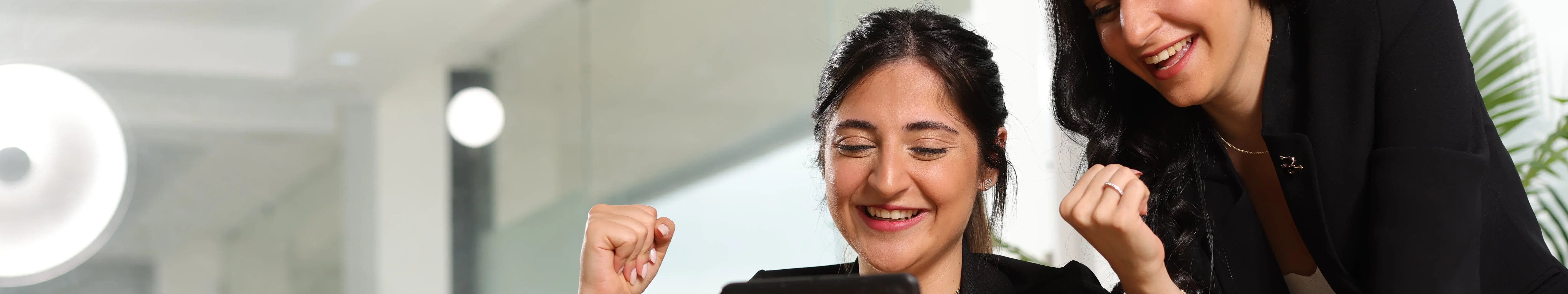 Deux femmes souriantes, l'une assise et l'autre debout à se côté, regardent le contenu d'une tablette tactile