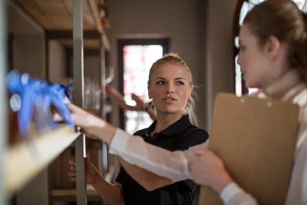 A woman in a storage room, inspecting inventory on shelves, holding a clipboard and engaging in a discussion.